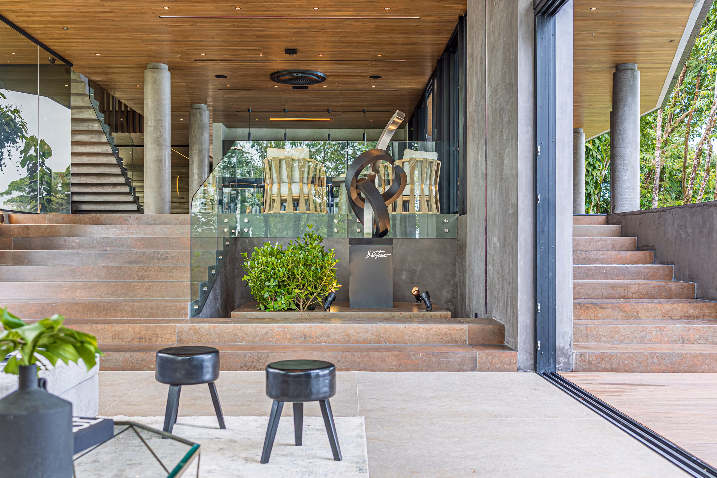 An open-riser concrete stair rising past full-height glazing with forest beyond.