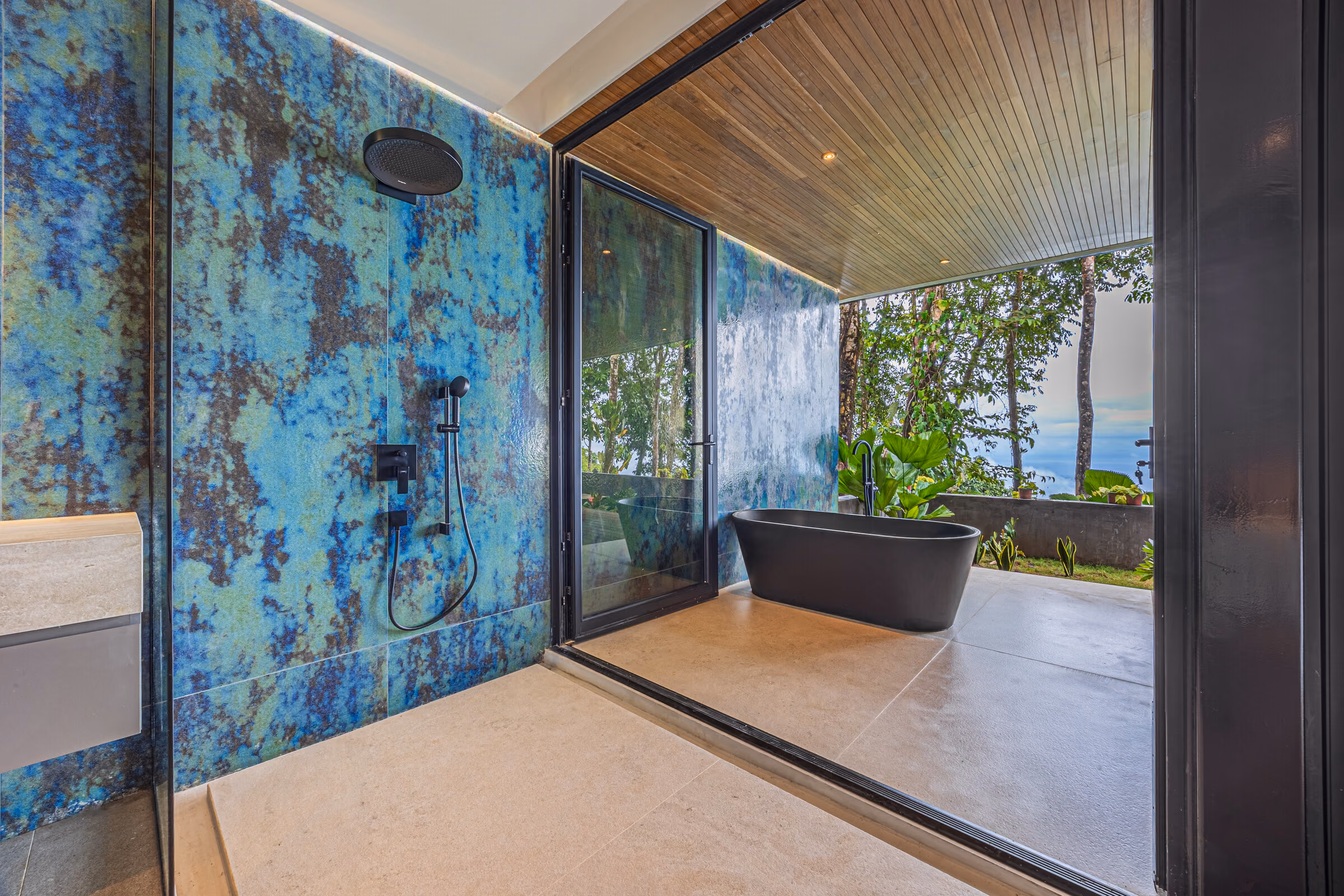 A bathroom finished in a large-format oxidised blue-green panel, with a freestanding dark tub and planting outside the glass.