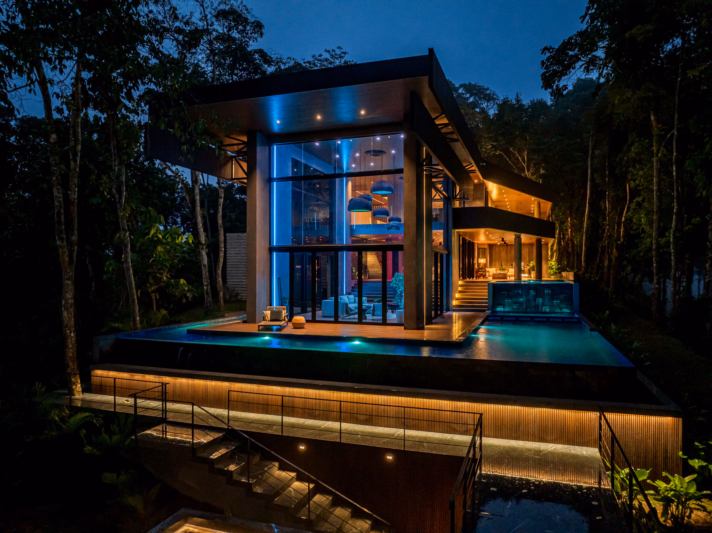 The residence at night with the lighting set to a cool teal scene, the pool lit and the forest dark around it.