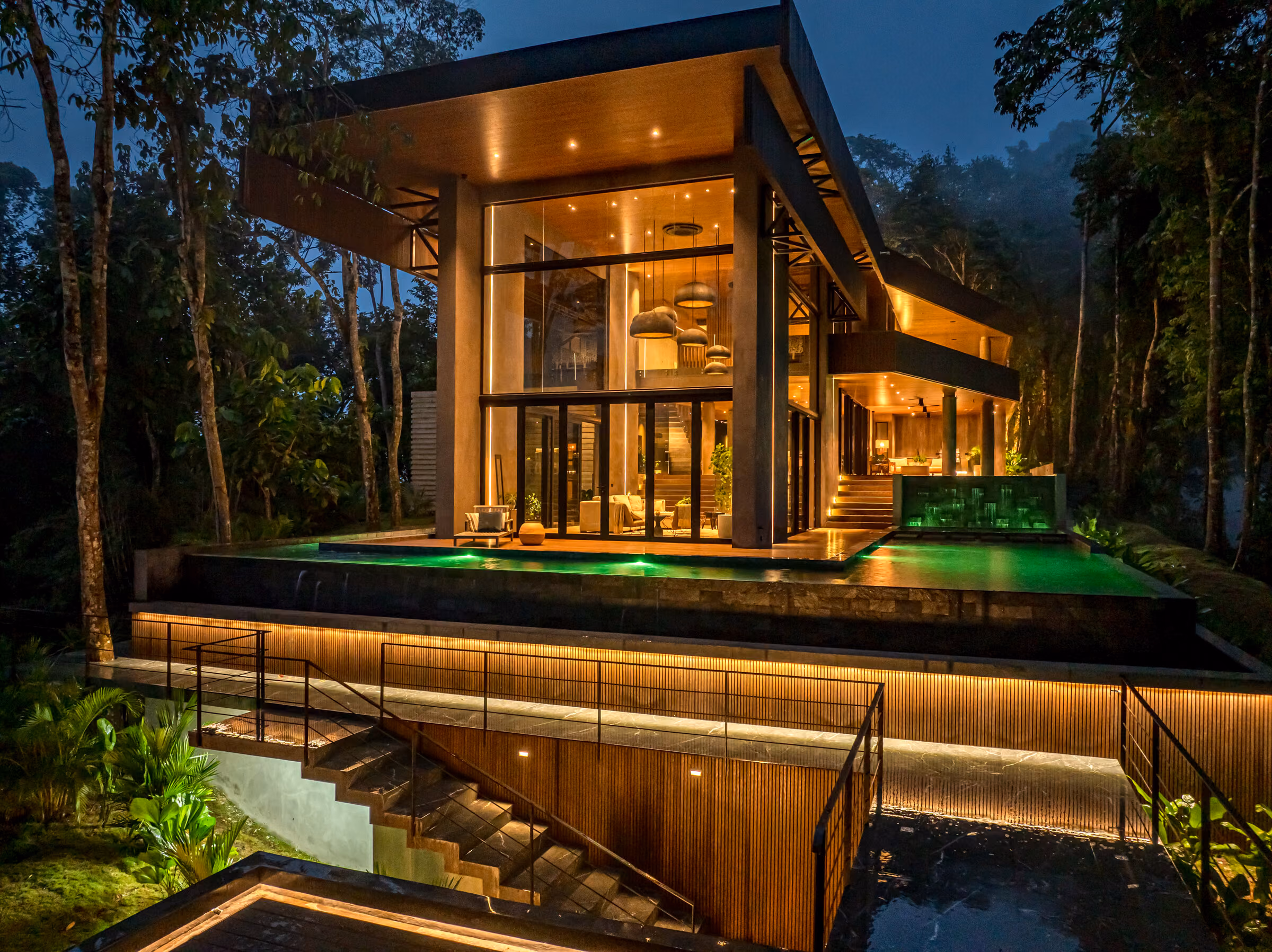 The residence at dusk, warm light in the timber-lined interior and the lit pool below.
