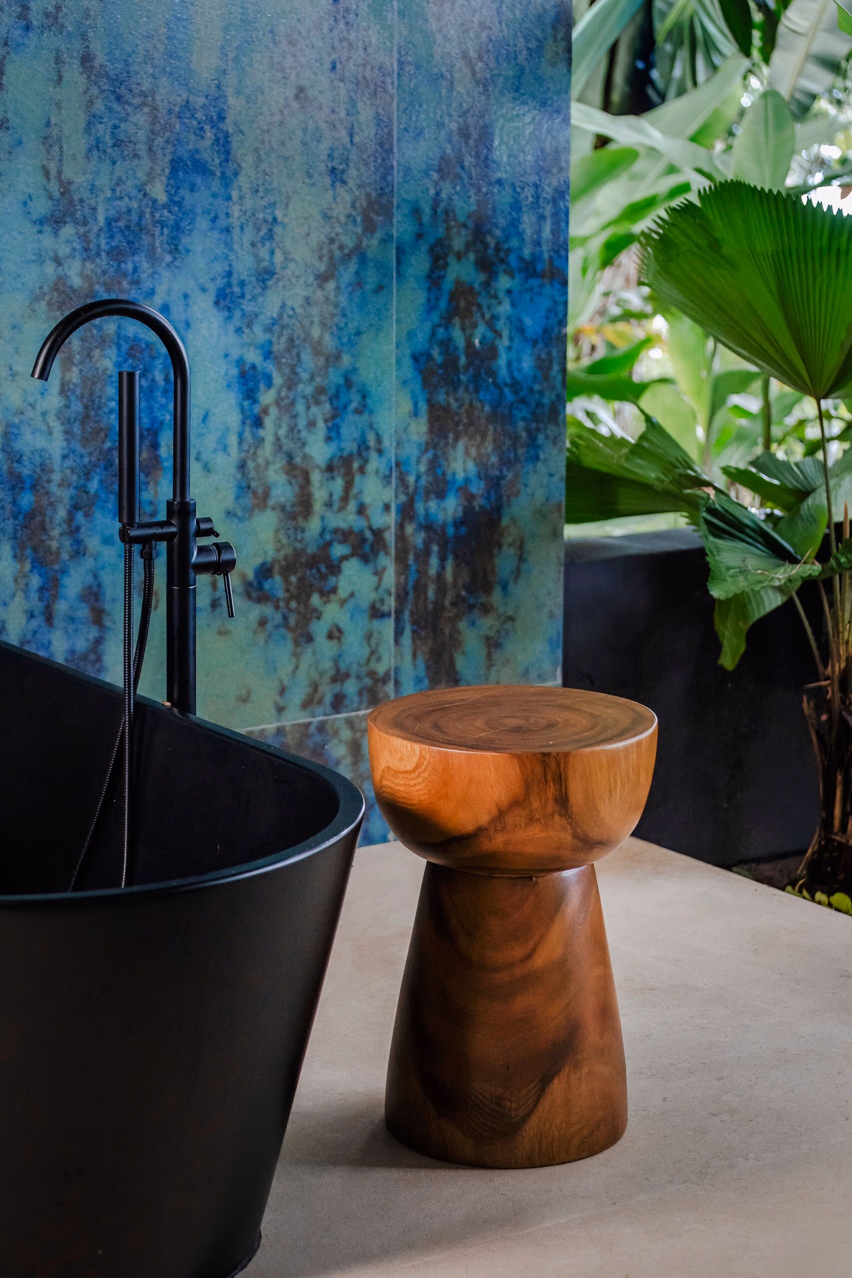 A turned timber stool beside a matt black freestanding bath, in front of an oxidised blue-green wall and forest glazing.