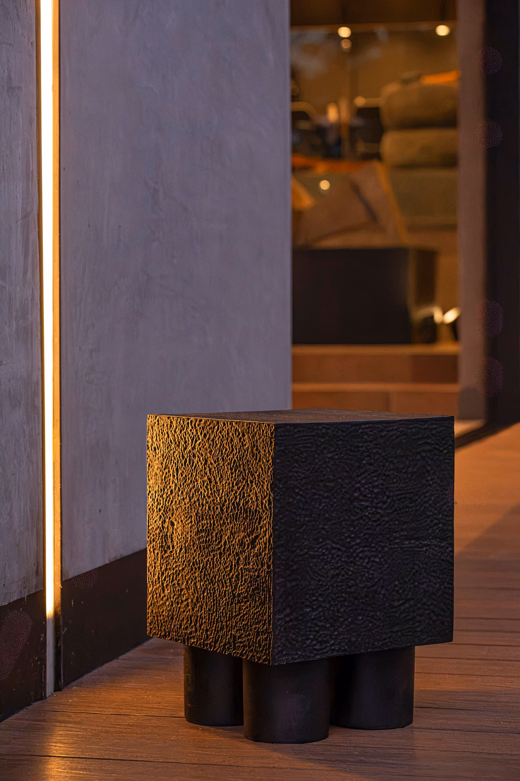 A black textured cube table on short round feet, catching a line of warm light on a timber floor.