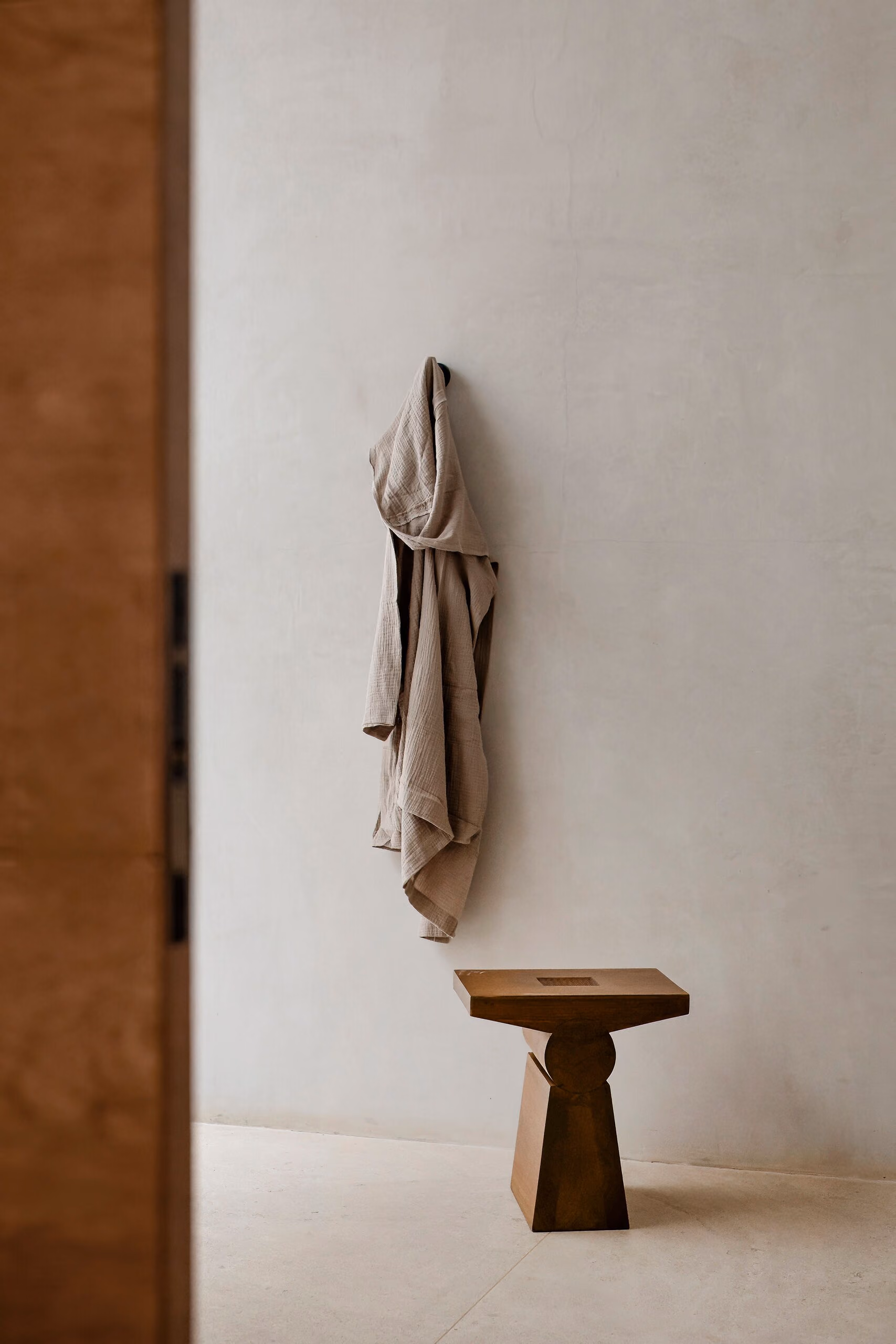 A small carved timber stool alone beneath a linen robe on a peg, against a pale plaster wall.