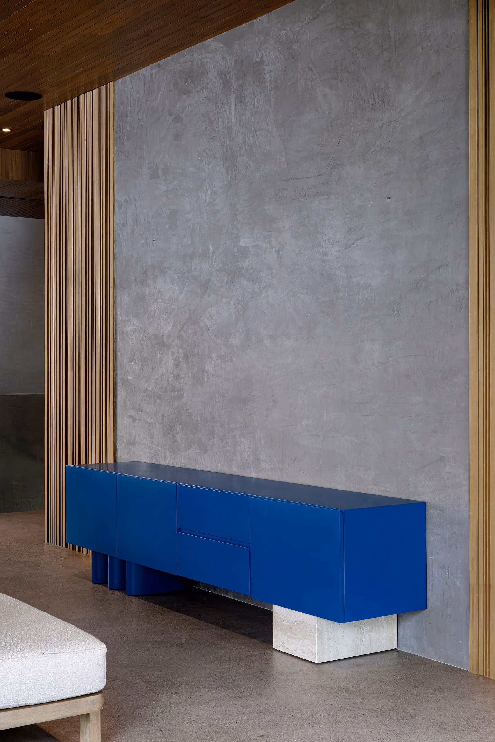 A long cobalt blue credenza on a pale plinth, set against grey concrete between vertical timber battens.