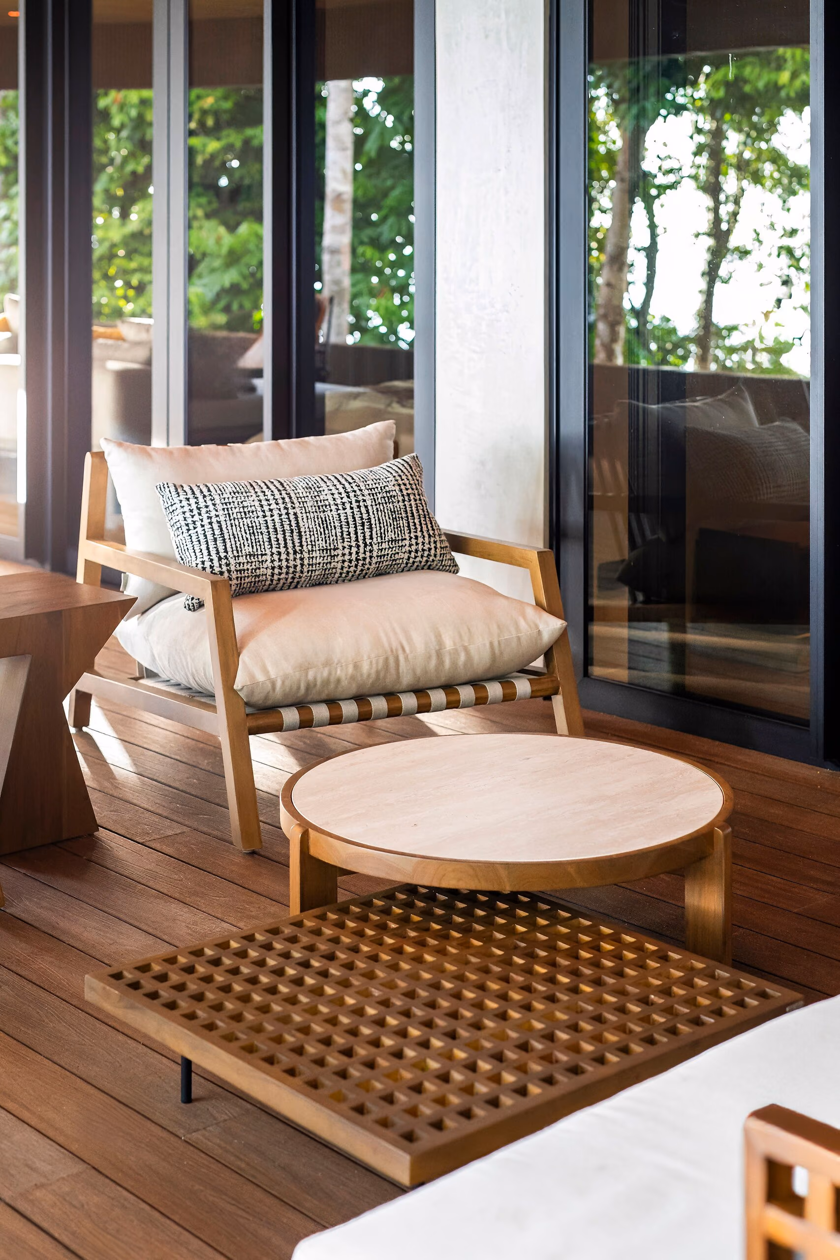 A timber armchair with a patterned cushion beside a round table on a woven lattice rug, on the covered deck.