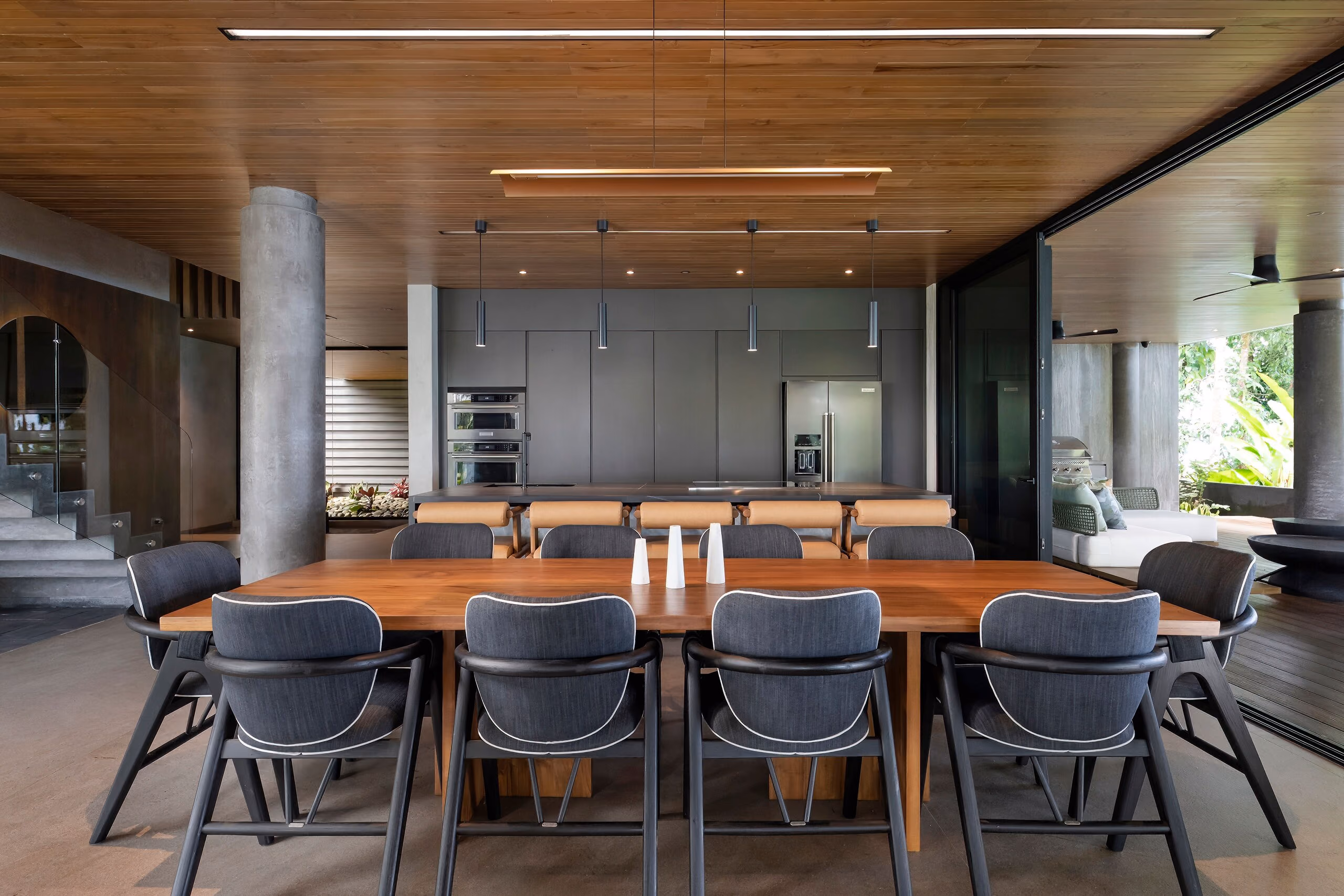 The dining room: a long timber table with dark upholstered chairs under a slatted teak ceiling, open to the kitchen beyond.