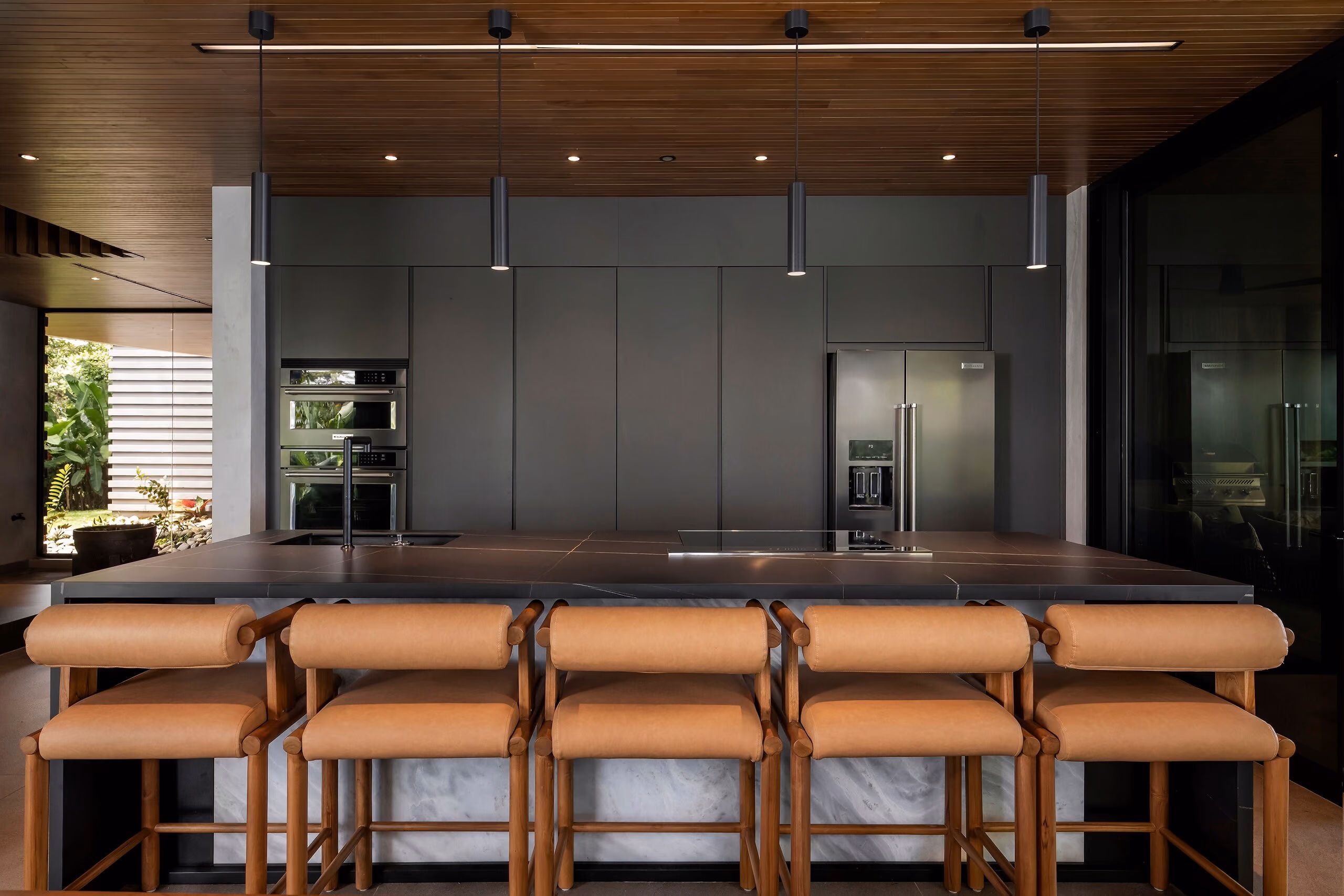 The kitchen island with a run of tan leather and timber bar stools, backed by dark cabinetry and integrated appliances.