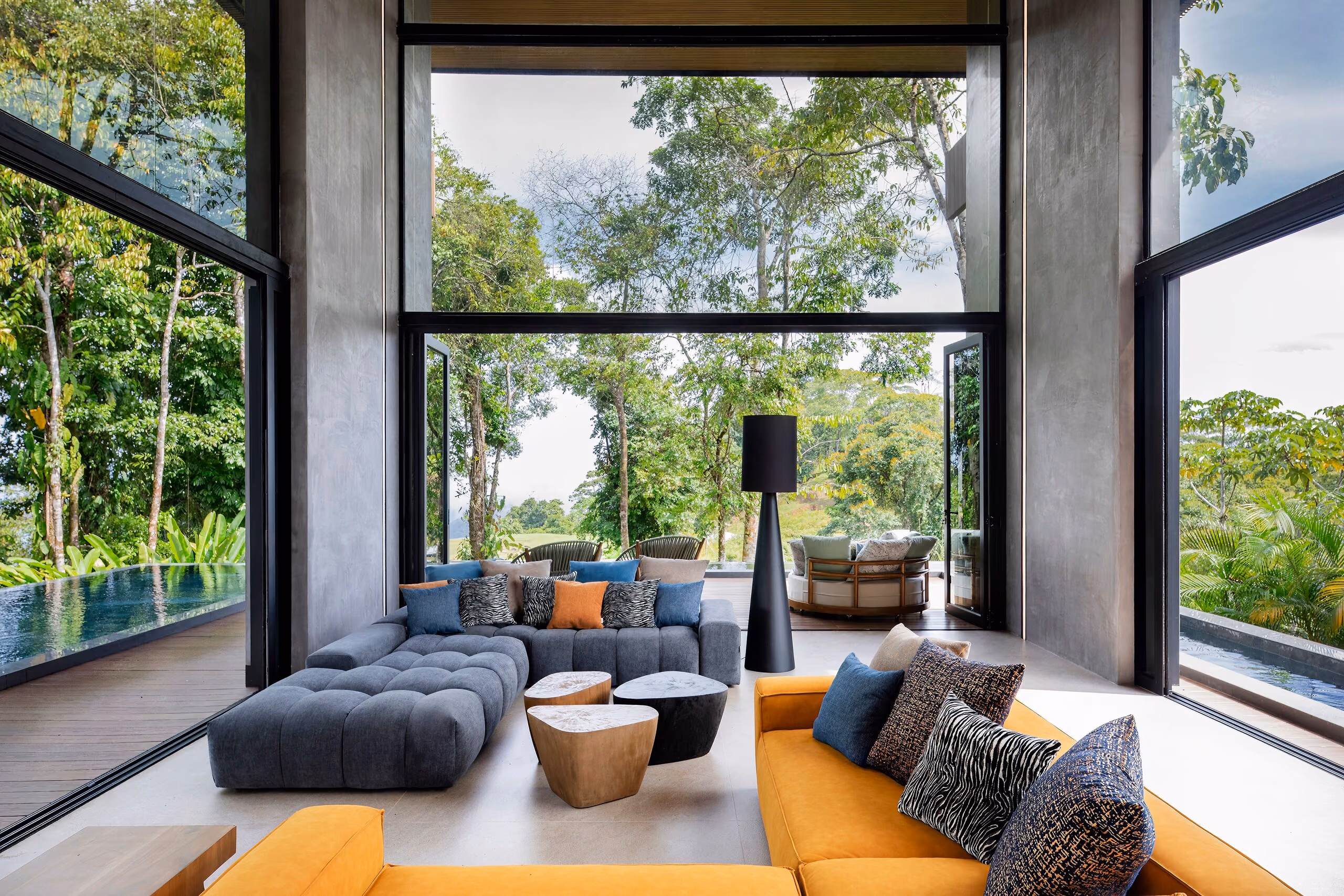 The main living room with its ochre sectional and grey tufted sofa facing full-height glazing onto the rainforest canopy.
