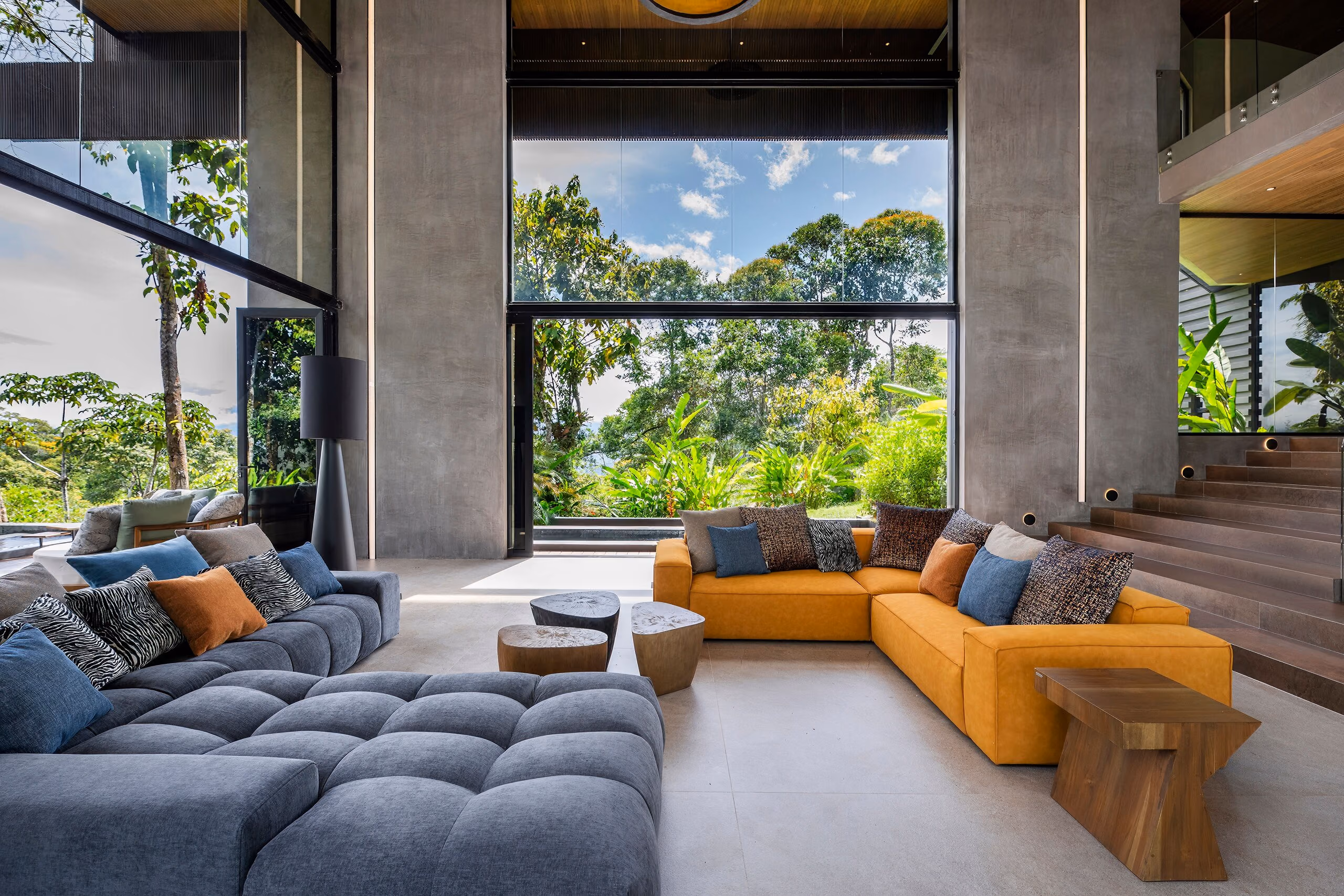 Wide view of the living room with the ochre sectional, blue and rust cushions and sculpted timber occasional tables, framed by tall windows.