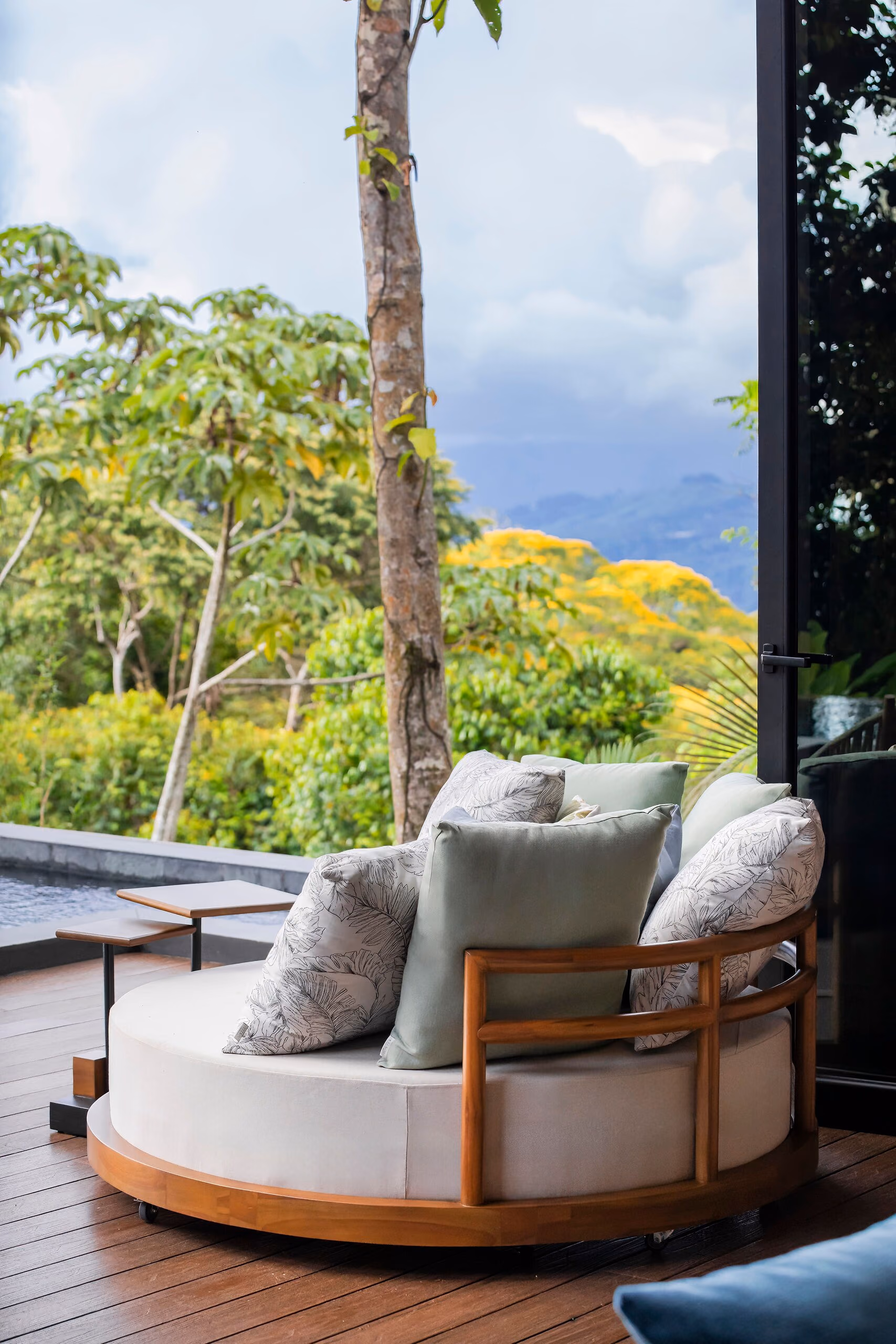 A circular timber and rope daybed with pale cushions on the deck, looking out over the treetops to the sky.
