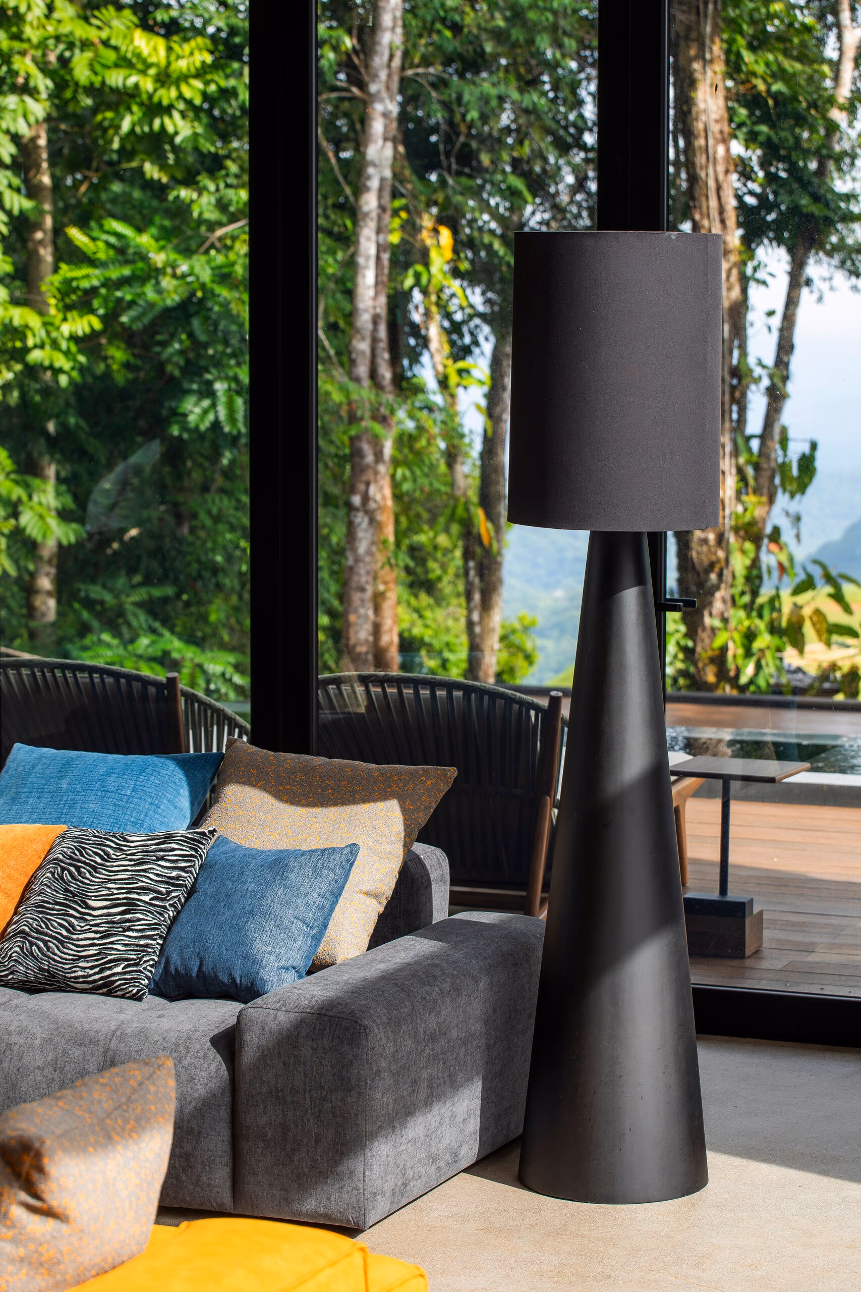 A tall black conical floor lamp beside a grey sofa dressed with ochre, teal and patterned cushions, with the canopy beyond.