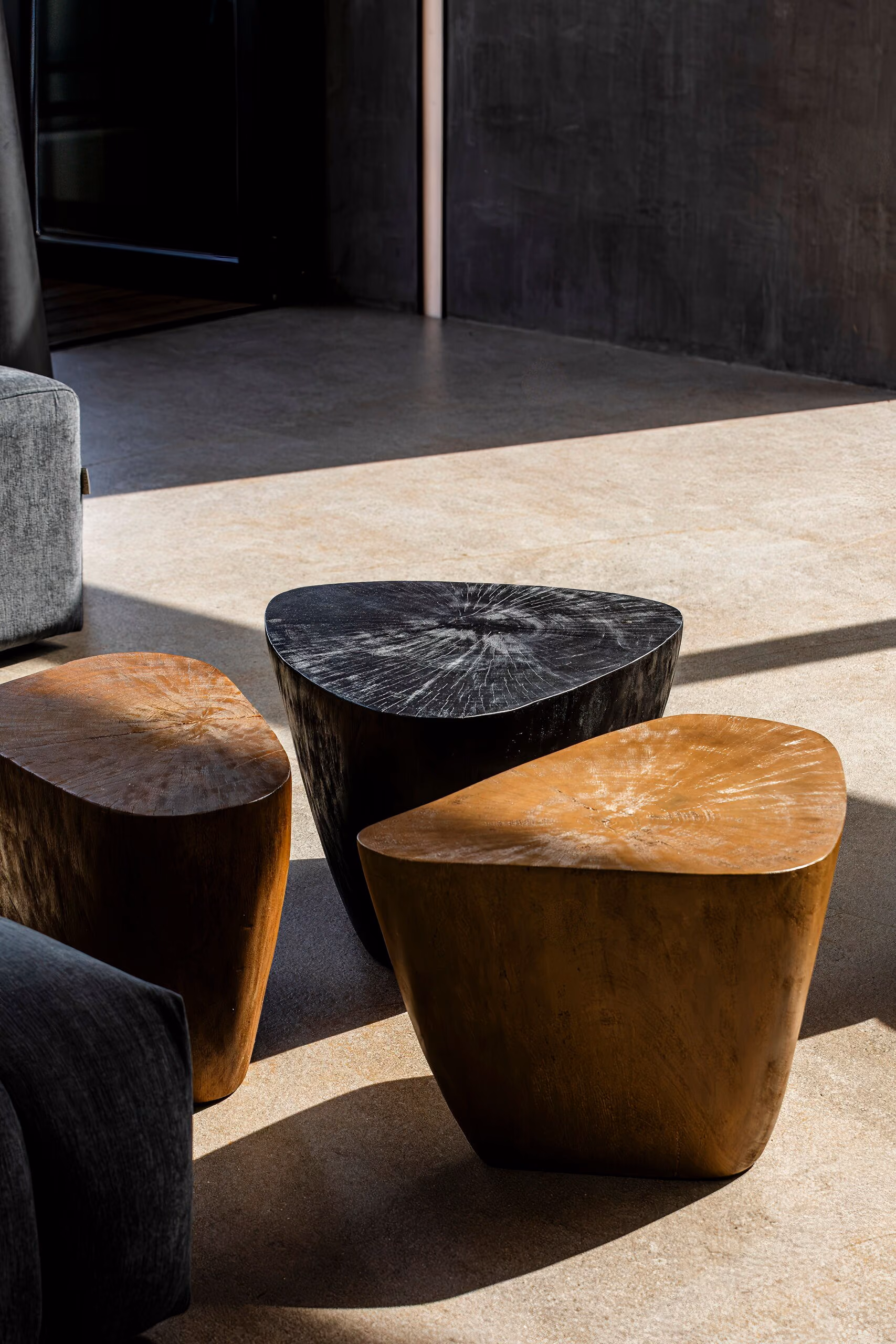 A black stone side table and two solid timber stump tables grouped on a pale floor in raking light, their end-grain and cut edges left visible.
