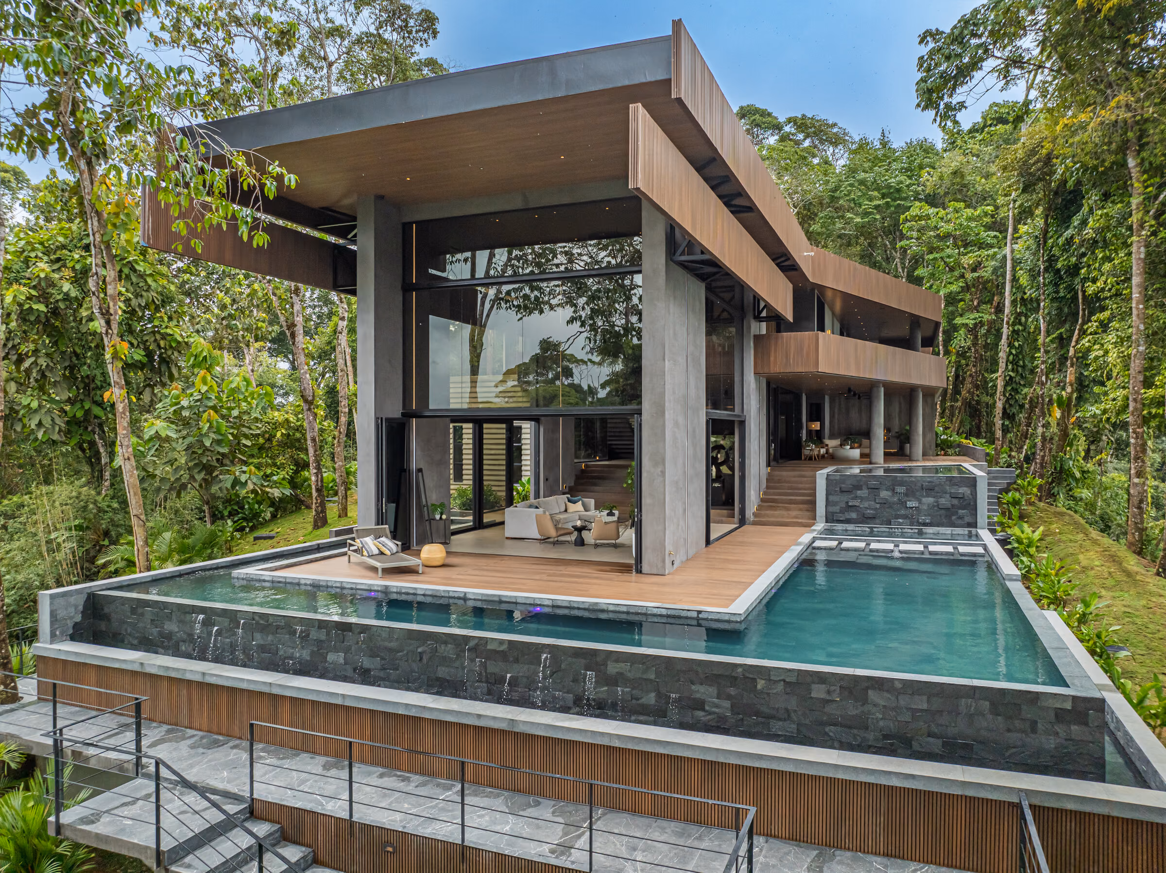 The residence viewed across its pool, dark concrete piers and deep roof overhangs against rainforest.