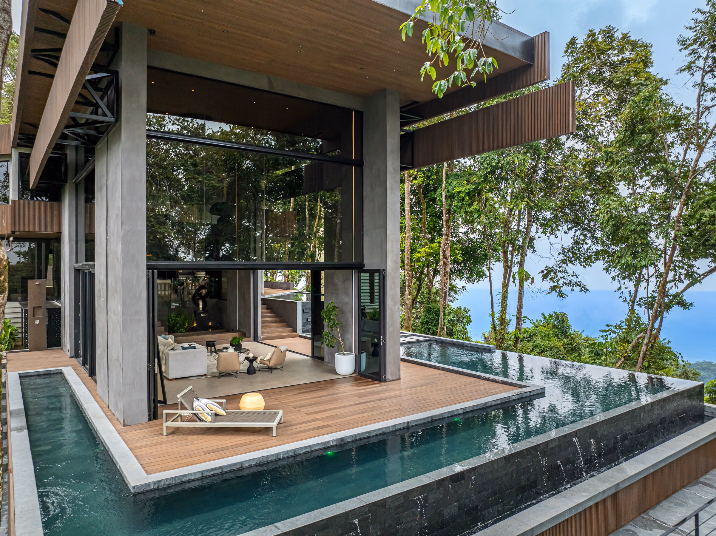 A shaded lounge terrace beside the pool, timber deck underfoot and rainforest rising on both sides.