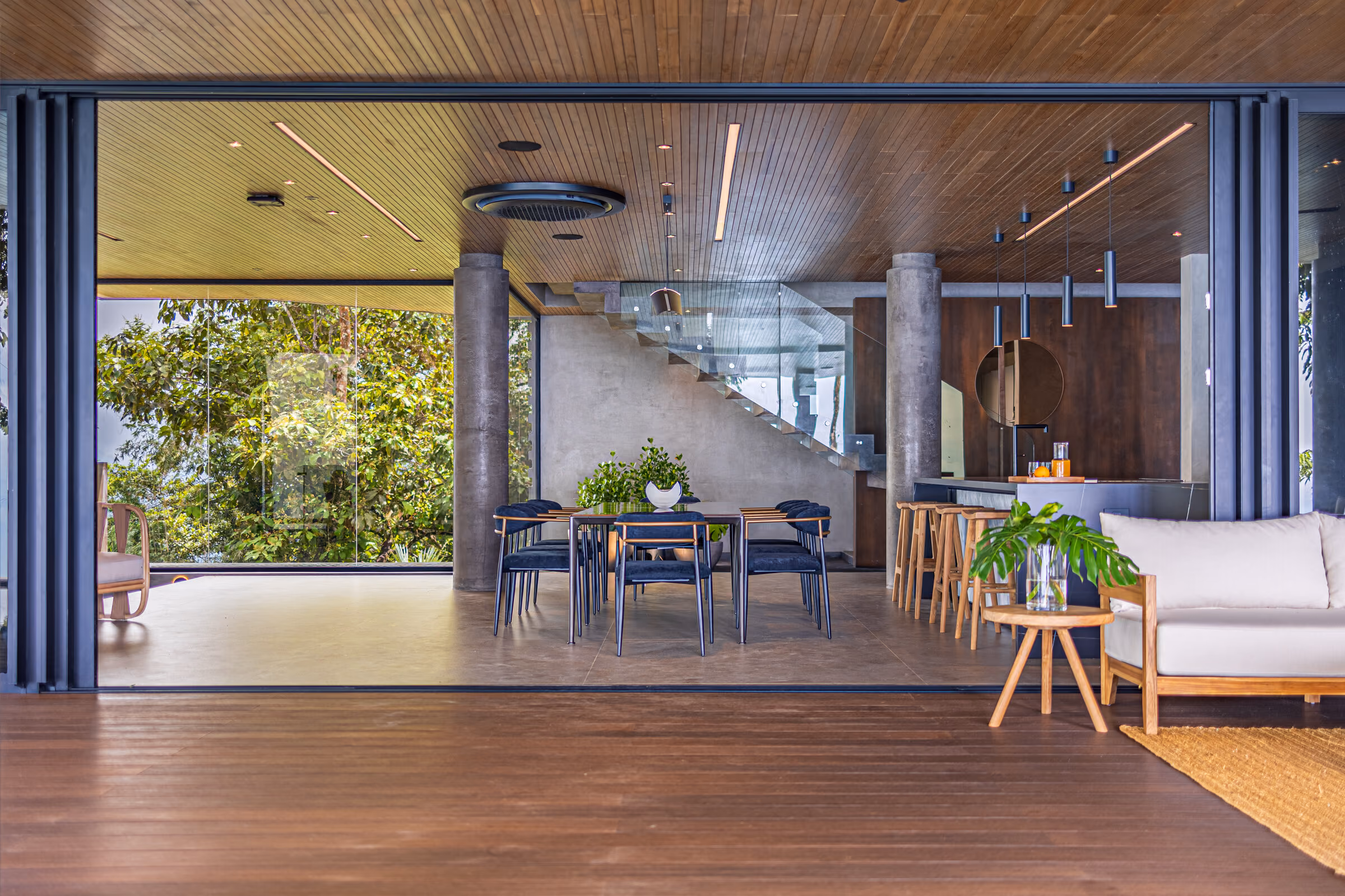 The principal living space: a triple-height room under a warm timber ceiling, open on two sides to terraces and forest, with the dining table and kitchen raised on the level above.